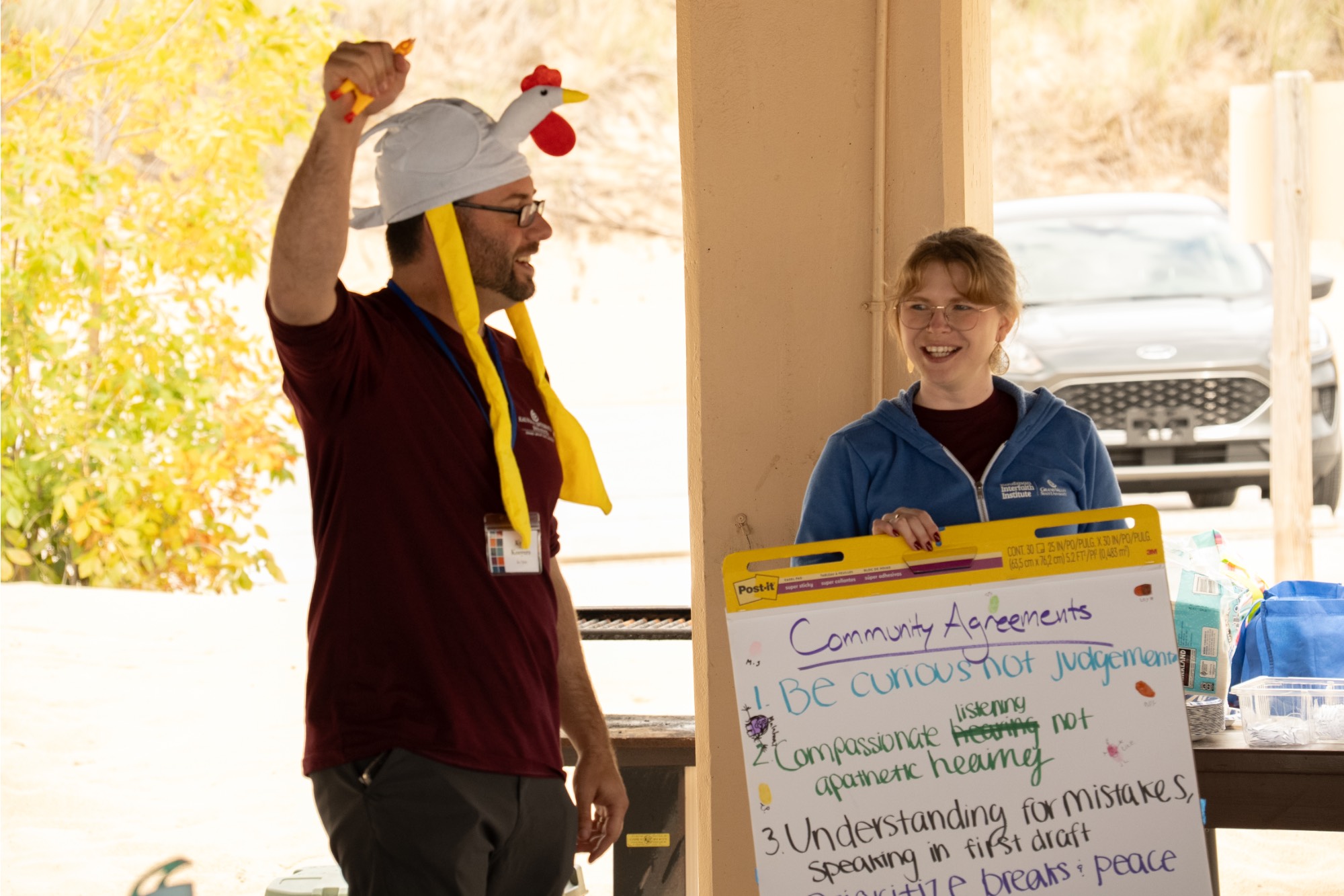 Man in a chicken hat squeaking a rubber chicken, woman laughing and holding a poster.
